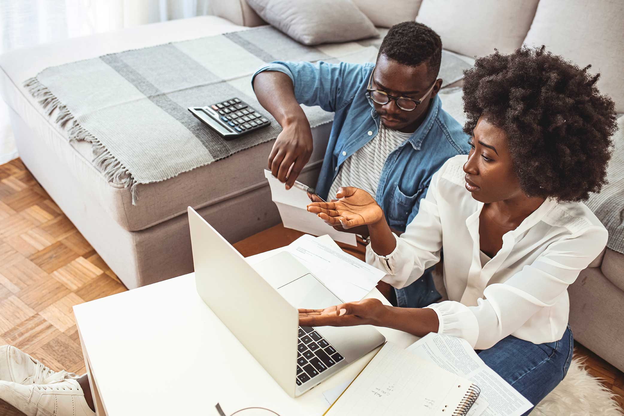 Image of a black man and black woman, working on a laptop computer. A calculator sits in the back ground. The couple looks upset, possibly victims of banking discrimination. shutterstock_1837384156